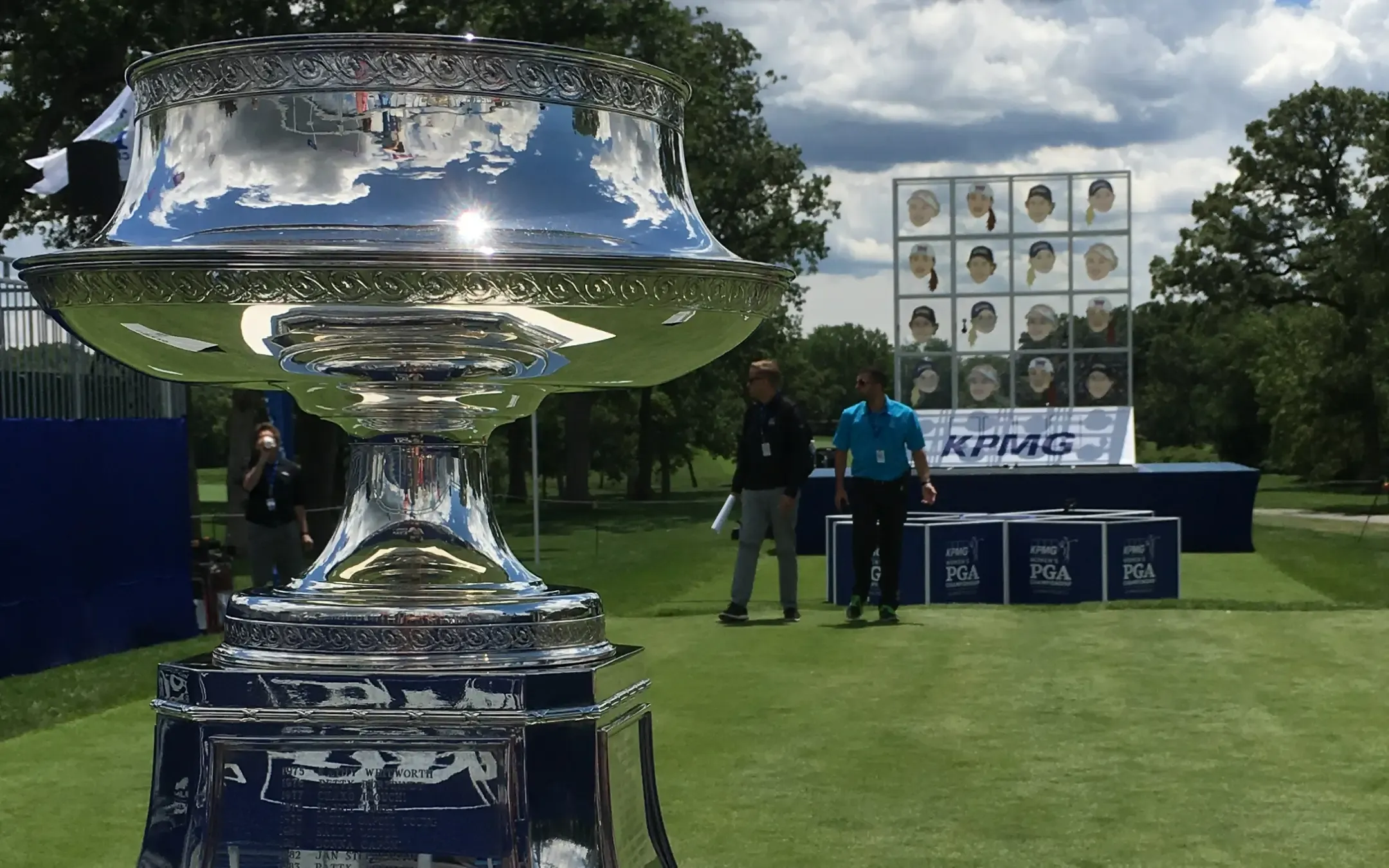 PGA trophy in foreground with reflective surface, event staff in background, and KPMG branding visible on display board.