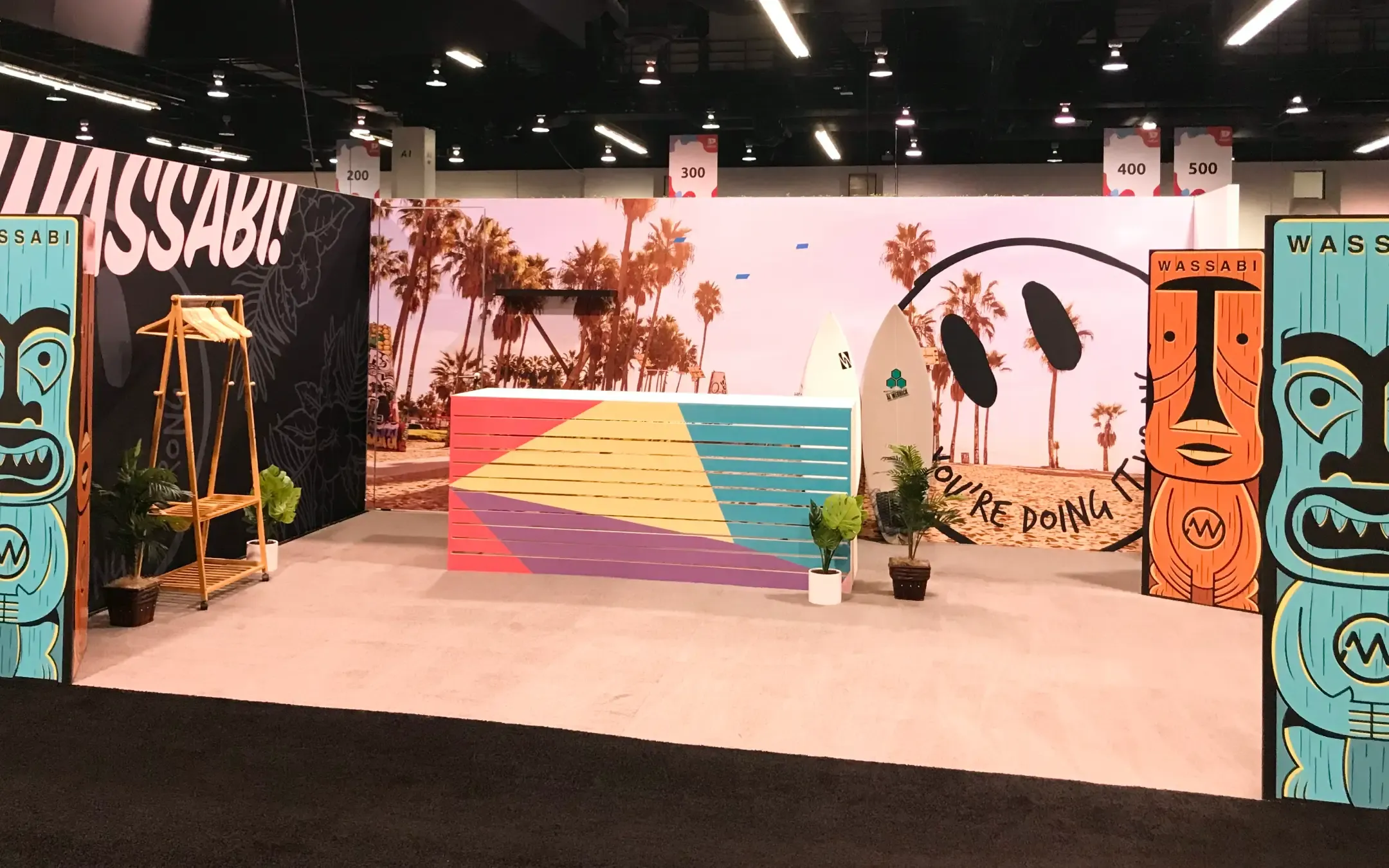 Colorful brand activation booth featuring WASSABI signage, vibrant surfboard designs, tropical background with palm trees, and engaging visual elements for experiential marketing.
